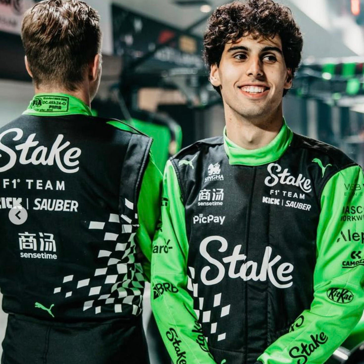 Gabriel And Nico wearing green and black racing suits with 'Stake' branding in a garage setting.