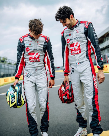 Oliver And Esteban race drivers in matching silver, black, and red Haas F1 racing suits walking on a racetrack, each holding a helmet. Visible branding includes MoneyGram and Haas.