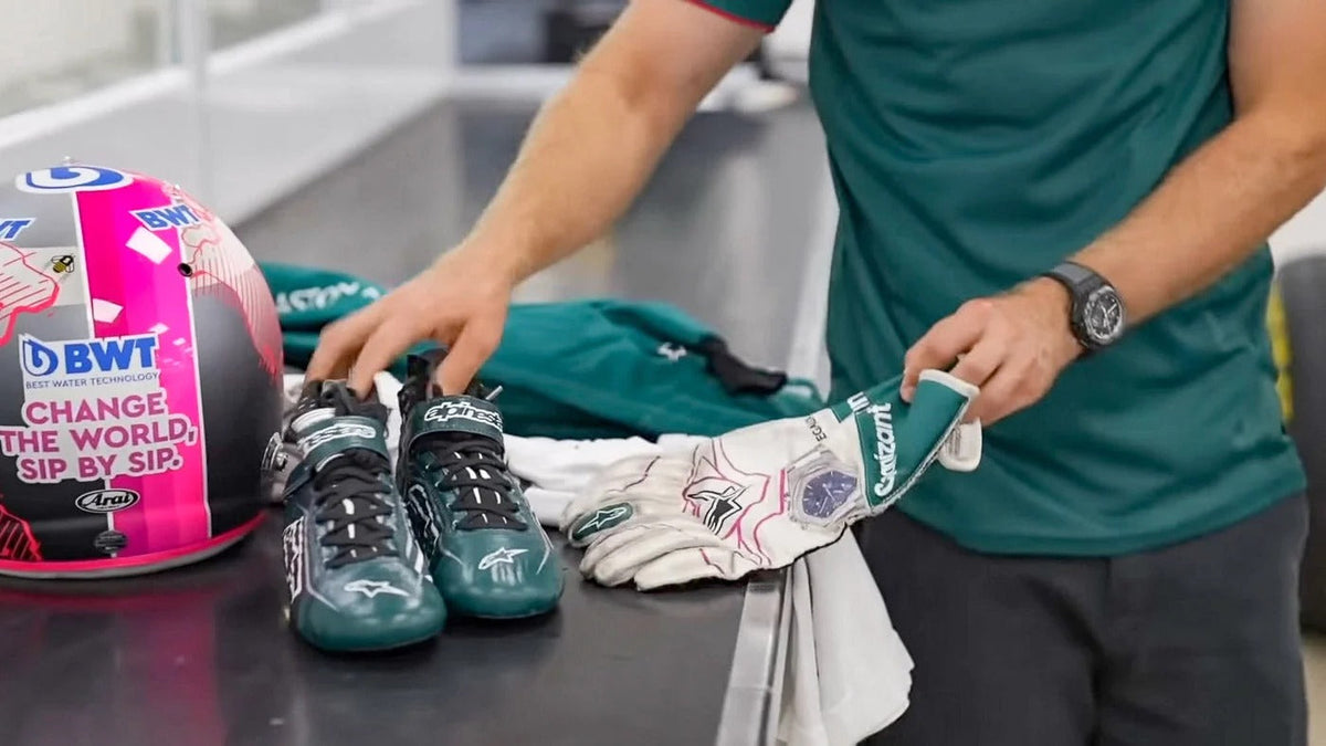 Lance Stroll in a teal shirt handles 2021 Replica Race Gloves F1 Aston Martin and teal racing boots on a table next to a pink and black racing helmet with visible sponsor logos.