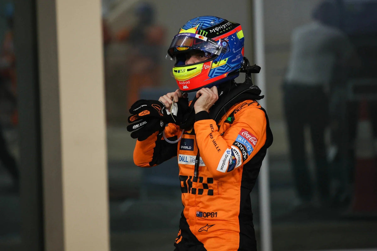 A race driver Oscar Piastri in an orange and black Alpinestars racing suit and a blue, red, and yellow helmet, adjusts their neck collar in a garage or pit area.