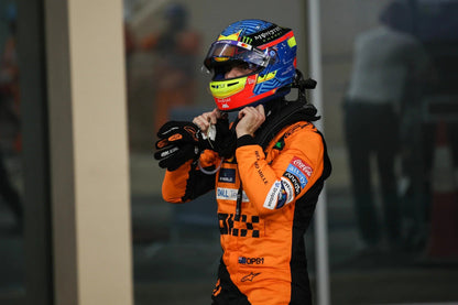 A race driver Oscar Piastri in an orange and black Alpinestars racing suit and a blue, red, and yellow helmet, adjusts their neck collar in a garage or pit area.
