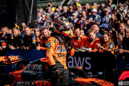 Oscar Piastri in orange racing suit and helmet standing next to a Formula 1 car with AWS branding, surrounded by a crowd.