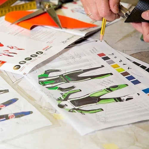 A close-up of a person's hands sketching a technical suit design on a sheet of paper, surrounded by color swatches, scissors, and orange paper on a worktable.